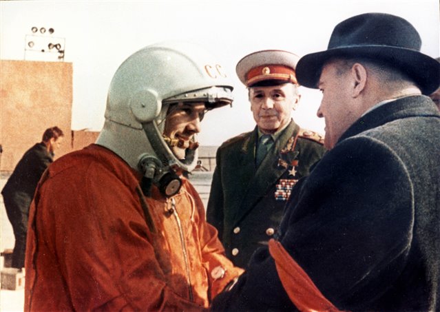 A man in an orange space suit talks to other men